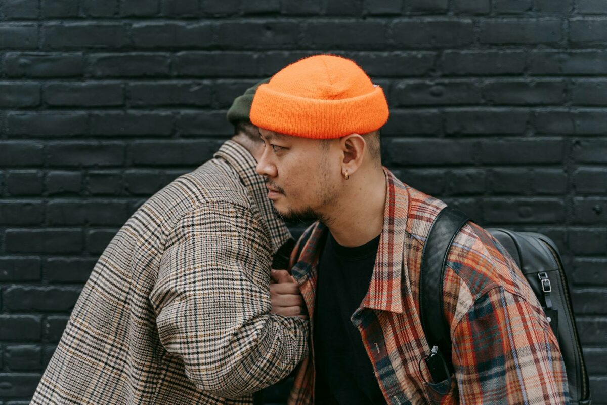 Two men wearing plaid jackets and beanies stand against a brick wall, portraying urban style.